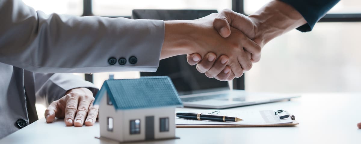 Deux personnes se serrent la main par-dessus une table où se trouvent une petite maquette de maison, un ordinateur portable et des documents.